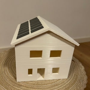 May include: A small, cream-colored model house with a pitched roof and rectangular windows. The roof features a series of dark gray solar panels. The house has a simple, modern design, and is likely a decorative item or architectural model.