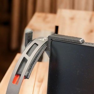 Könnte beinhalten: Ein schwarzer und grauer Kopfhörerhalter, der an einem schwarzen Monitor befestigt ist. Der Kopfhörerhalter besteht aus einem strukturierten, dunkelgrauen Material und hat ein gebogenes Design, um den Kopfhörer zu halten. Der Kopfhörer selbst ist schwarz und grau mit einem roten Akzent.