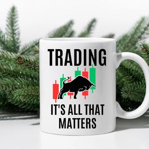 Può includere: Tazza in ceramica bianca con la scritta "TRADING" sopra la frase "IT'S ALL THAT MATTERS". La tazza presenta una grafica di toro nero e grafici a candela rossi e verdi del mercato azionario. La tazza ha un manico bianco.