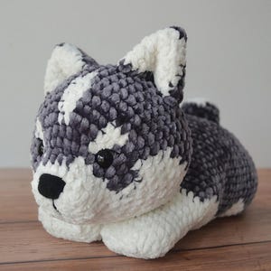Puede incluir: Un juguete de perro husky hecho a mano. El juguete presenta un hilo suave y texturizado gris y blanco. El husky tiene ojos negros y una nariz negra. Las orejas y las patas son blancas. El juguete está sobre una superficie de madera.