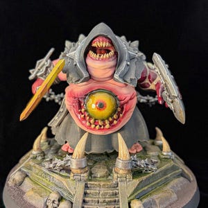 May include: A detailed miniature figure of a grotesque creature with a large eye, sharp teeth, and a hooded cloak. It holds a sword and shield, standing on a base with skulls and a stone structure. The figure is painted in shades of pink, gray, and yellow.