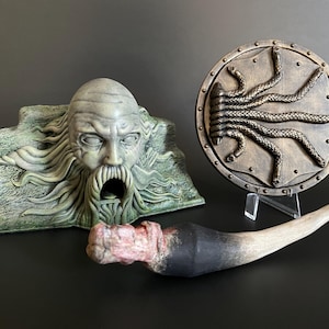 Puede incluir: Un conjunto de tres accesorios de temática fantástica, que incluye una cabeza esculpida de color verde y marrón con una boca abierta, un escudo redondo con un diseño de serpiente dorado y marrón, y un cuerno largo, marrón y negro.