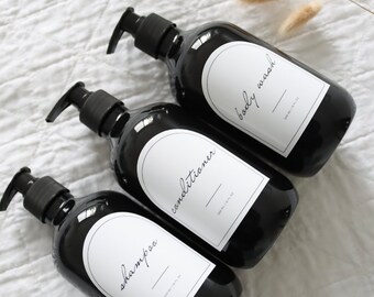 May include: Three black pump bottles labeled "shampoo", "conditioner", and "body wash". Each bottle has a white label with black text. The bottles are arranged on a white, textured surface. The bottles are 16 fl oz.