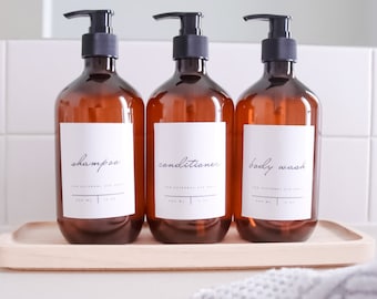 May include: Three amber-colored pump bottles labeled "shampoo", "conditioner", and "body wash" with black pumps and white labels. The bottles sit on a light wooden tray. Each bottle has a capacity of 500 ml / 16 oz.