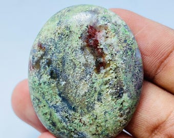 May include: Oval-shaped gemstone with a mottled surface. The stone features a mix of green, brown, and red hues, with a textured appearance. The gemstone is held in a hand, showcasing its natural colors and patterns.