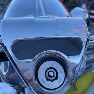 May include: Close-up of a chrome motorcycle ignition switch. The switch is labeled "UNLOCK" and "LOCK". The surrounding area is also chrome, with a black textured pad. The top of the motorcycle is visible in the background.