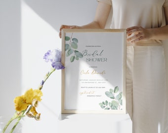 May include: A white card with green eucalyptus leaves and gold lettering. The card is a bridal shower invitation for Cule Deveda. The invitation includes the date, time, location, and RSVP information. The card is framed in a light wood frame.