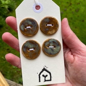 May include: Four round ceramic buttons with a glossy brown and teal glaze, each featuring two holes with white thread, attached to a rectangular white display card that has a small black house-shaped logo printed at the bottom centre.
