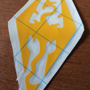 May include: A yellow vinyl decal featuring the emblem of the video game The Elder Scrolls V: Skyrim. The emblem is a dragon silhouette within a shield shape. A blue grid pattern is visible on the backing.