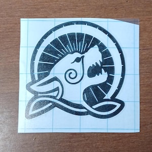 Puede incluir: Adhesivo de vinilo en blanco y negro con la cabeza estilizada de un lobo dentro de un diseño circular. El lobo tiene la boca abierta con dientes visibles, y el diseño incluye remolinos y líneas decorativas. La pegatina está sobre un respaldo cuadriculado.