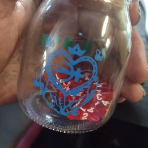 May include: A glass jar with a blue and white design of a heart with a crown and wings. The jar contains several red dice with white numbers.