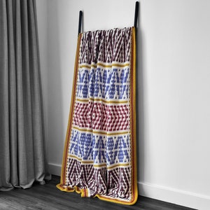 Peut inclure: Une couverture au crochet colorée avec un motif géométrique dans des tons de bleu, rouge, jaune et blanc. La couverture est accrochée à un crochet contre un mur blanc. Modèle de crochet téléchargeable en PDF.