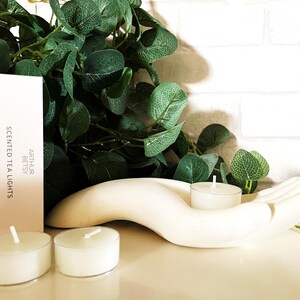 Op de afbeelding: Een witte, handvormige kaarsenhouder met twee brandende waxinelichtjes. Een doosje met "Scented Tea Lights" staat naast de houder. Groene bladeren zijn op de achtergrond. De waxinelichtjes zijn klein, rond en wit.