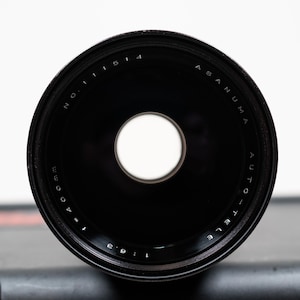 May include: A black Asanuma Auto-Tele lens with a central aperture. The lens has white text that reads "NO. 11514" and "f=400mm 1:6.3". The lens is set against a dark gray case.