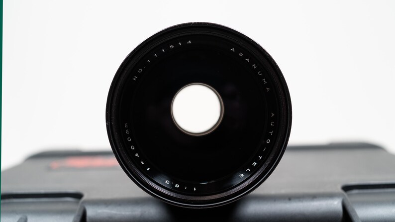 May include: A black Asanuma Auto-Tele lens with a central aperture. The lens has white text that reads "NO. 11514" and "f=400mm 1:6.3". The lens is set against a dark gray case.