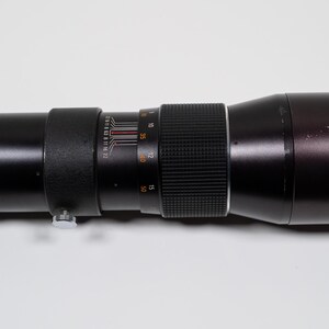 May include: A black telephoto camera lens with silver and orange markings. The lens has multiple rings and a textured grip. The lens is cylindrical and appears to be made of metal and glass.