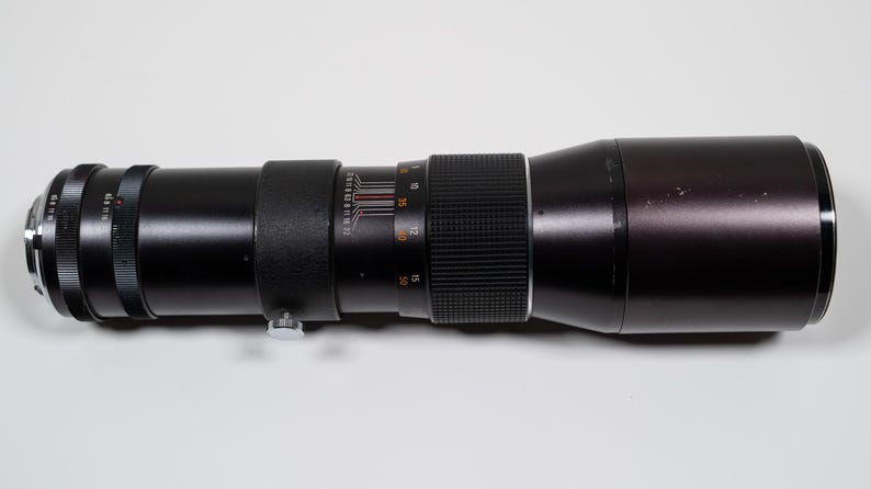 May include: A black telephoto camera lens with silver and orange markings. The lens has multiple rings and a textured grip. The lens is cylindrical and appears to be made of metal and glass.