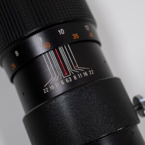 May include: Close-up of a black camera lens with white and red markings. The lens has a textured grip and a silver adjustment knob. Numbers and lines indicate focal length and aperture settings.