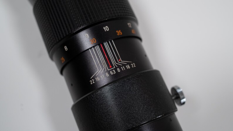 May include: Close-up of a black camera lens with white and red markings. The lens has a textured grip and a silver adjustment knob. Numbers and lines indicate focal length and aperture settings.