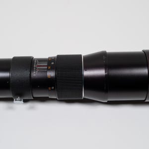 May include: A long, black camera lens with silver and black details. The lens has a ribbed focus ring and measurement markings. The lens is designed for photography and is likely used for capturing distant subjects.