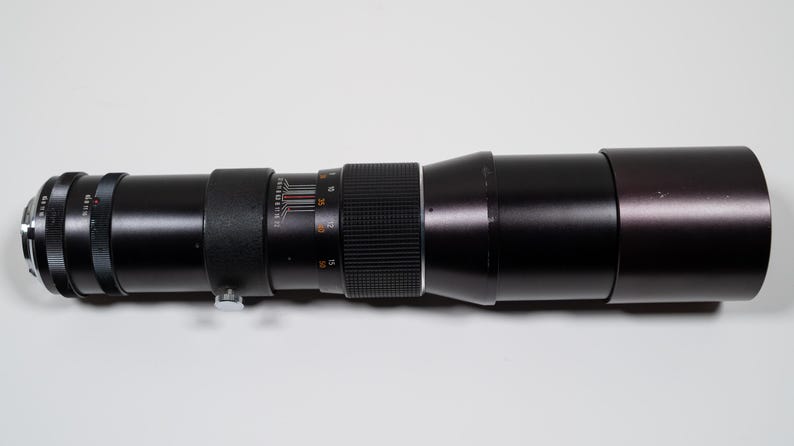May include: A long, black camera lens with silver and black details. The lens has a ribbed focus ring and measurement markings. The lens is designed for photography and is likely used for capturing distant subjects.