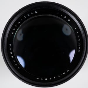 May include: A black Asanuma Auto-Tele camera lens with a 400mm focal length and f/6.3 aperture. The lens has white text that reads "ASANUMA AUTO-TELE" and "1:6.3 f=400mm". The lens is set against a white background.