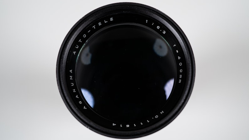 May include: A black Asanuma Auto-Tele camera lens with a 400mm focal length and f/6.3 aperture. The lens has white text that reads "ASANUMA AUTO-TELE" and "1:6.3 f=400mm". The lens is set against a white background.