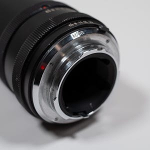 May include: Close-up of a black camera lens with a silver-colored mount. The lens has a ridged design and a red dot. The mount has the letters "M/SR" engraved on it. The lens is on a white surface.