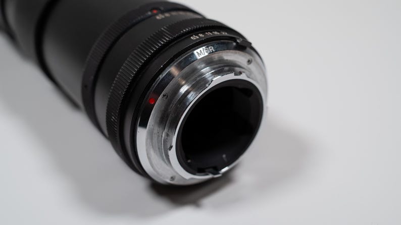 May include: Close-up of a black camera lens with a silver-colored mount. The lens has a ridged design and a red dot. The mount has the letters "M/SR" engraved on it. The lens is on a white surface.