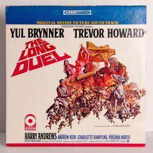 May include: A vintage record album cover featuring a colorful illustration of a train and soldiers. The album title is "The Long Duel" and the actors Yul Brynner and Trevor Howard are listed. The album is a Paramount Pictures release.