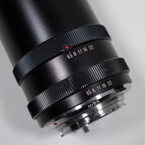 May include: Close-up of a black camera lens with focus settings. The lens has a ridged focus ring with white numbers and a red dot. The lens is angled, revealing the M/SR mount.