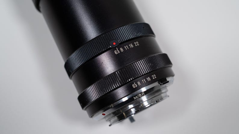 May include: Close-up of a black camera lens with focus settings. The lens has a ridged focus ring with white numbers and a red dot. The lens is angled, revealing the M/SR mount.