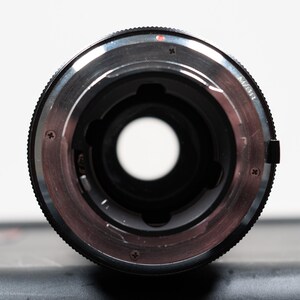 May include: Close-up of a camera lens mount. The lens has a black exterior with a silver-colored interior ring. The lens has a circular opening in the center. The lens is on a dark surface with a white background.