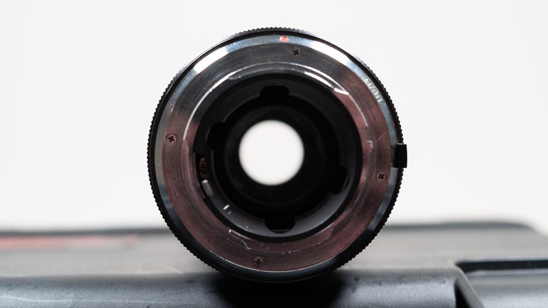 May include: Close-up of a camera lens mount. The lens has a black exterior with a silver-colored interior ring. The lens has a circular opening in the center. The lens is on a dark surface with a white background.