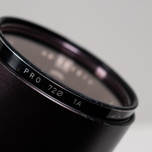 May include: Close-up of a black camera lens filter with the text "PRO 720 1A" in white. The filter has a dark, reflective surface and is set against a light gray background. The lens is cylindrical.
