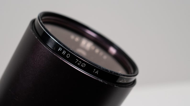 May include: Close-up of a black camera lens filter with the text "PRO 720 1A" in white. The filter has a dark, reflective surface and is set against a light gray background. The lens is cylindrical.