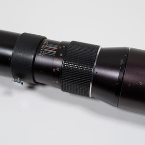 May include: A black telephoto camera lens with a ribbed focus ring and measurement markings. The lens has a silver adjustment ring and a long, cylindrical body. The lens is lying on a white surface.