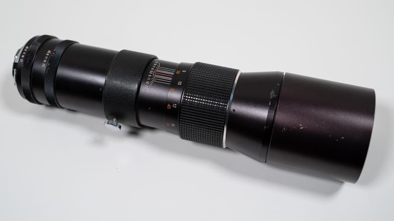 May include: A black telephoto camera lens with a ribbed focus ring and measurement markings. The lens has a silver adjustment ring and a long, cylindrical body. The lens is lying on a white surface.
