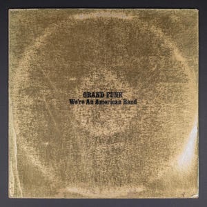 May include: A vintage album cover with a gold-toned, textured surface. The text "GRAND FUNK" and "We're An American Band" are printed in black, bold font. The album cover has a worn appearance, suggesting it is a used or collectible item.