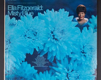 Könnte beinhalten: Albumcover für Ella Fitzgeralds "Misty Blue". Das Cover zeigt ein blaues Blumendesign mit dem Albumtitel und dem Namen des Künstlers in weißem Text. Ein Porträt von Ella Fitzgerald befindet sich in der oberen rechten Ecke. Das Capitol Records-Logo ist sichtbar.