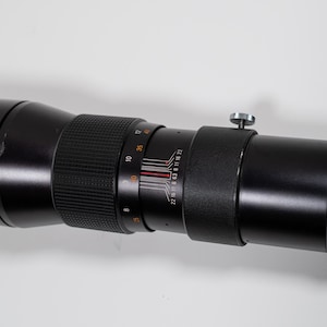 May include: A black telephoto camera lens with various adjustment rings and a silver knob. The lens has a ribbed grip section and measurement markings. The lens is designed for capturing distant subjects with precision.