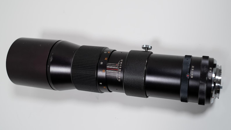 May include: A black telephoto camera lens with various adjustment rings and a silver knob. The lens has a ribbed grip section and measurement markings. The lens is designed for capturing distant subjects with precision.