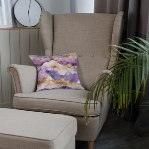 Op de afbeelding: Een beige fauteuil met een kussen met een paars en gouden bergpatroon. De stoel staat naast een beige voetenbank en een groene plant in een pot.