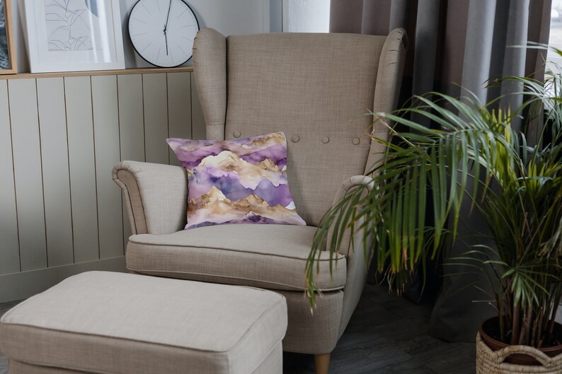 Op de afbeelding: Een beige fauteuil met een kussen met een paars en gouden bergpatroon. De stoel staat naast een beige voetenbank en een groene plant in een pot.