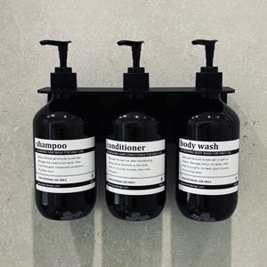 Pode incluir: Três garrafas pretas com bomba e etiquetas brancas montadas em uma prateleira de metal preta. Os rótulos dizem "shampoo", "condicionador" e "gel de banho". Cada garrafa tem um rótulo branco com texto preto que descreve o produto e as instruções de uso.