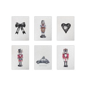 Könnte beinhalten: Set aus sechs rechteckigen Kunstdrucken mit Aquarellillustrationen. Die Drucke zeigen eine schwarze Schleife, ein Herz, ein Auto mit Gepäck und Nussknackersoldaten in roten und blauen Uniformen. Der Hintergrund ist ein gesprenkeltes Off-White.
