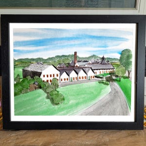 May include: Watercolor painting of a distillery with white buildings, green grass, and a blue sky. The artwork is framed in a black frame and displayed on a wooden surface. A bottle of liquor is visible on the left.