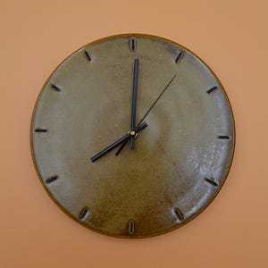 Op de afbeelding: Een bruine keramische klok met zwarte wijzers en gouden accenten. De wijzerplaat van de klok heeft een subtiele, getextureerde afwerking.