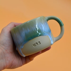Op de afbeelding: Een keramische mok met een matte beige basis en een glanzend groen en blauw druipglazuur op het bovenste gedeelte. Het woord URS is in het onderste beige gedeelte gestempeld. De mok heeft een gebogen handvat en een gladde, afgeronde drinkrand.