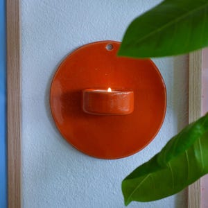 May include: A circular, glossy orange ceramic wall sconce featuring a small, integrated cup-shaped holder at the center. A lit tea light candle sits inside the holder, emitting a warm glow. A small hole is visible at the top for mounting.